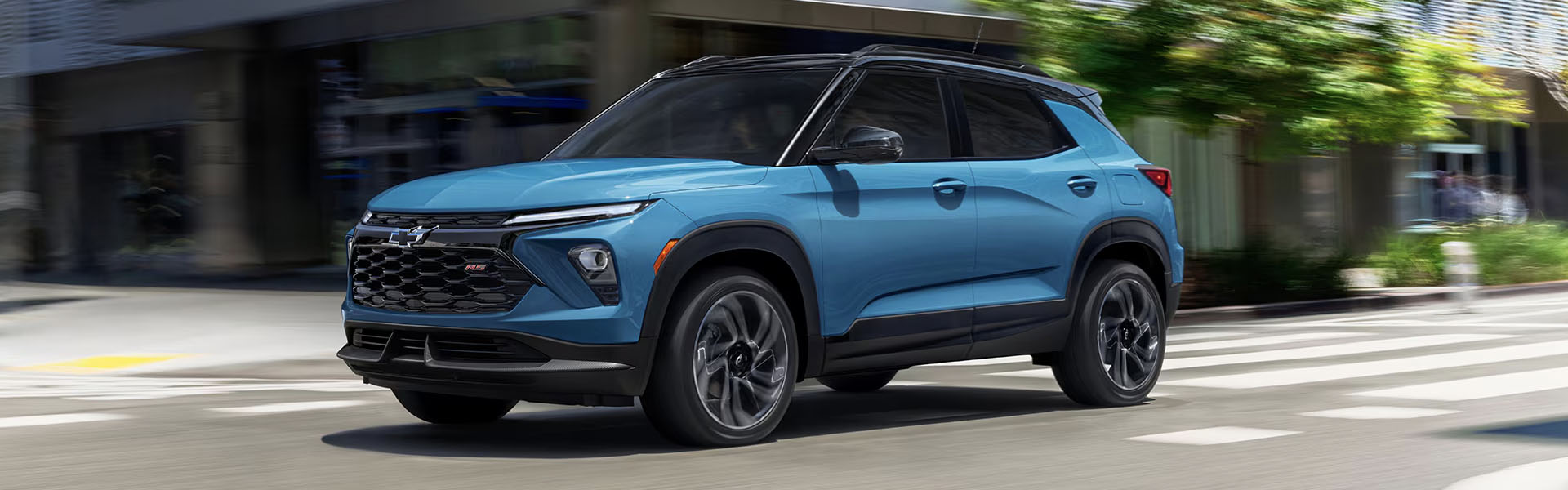 2025 Chevrolet Trailblazer compact SUV in blue driving through a modern city street, showcasing updated exterior design, sporty grille, LED headlights, alloy wheels, and urban crossover styling.