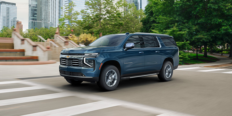 A large blue Suburban drives through a city intersection with modern buildings and lush greenery in the background, conveying a sense of strength and adventure.