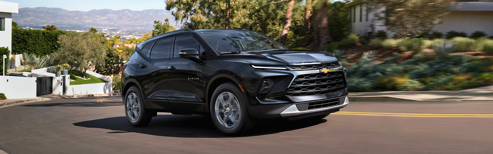 2026 Chevrolet Blazer RS, a black SUV, driving on a winding road with lush greenery and upscale homes in the background.