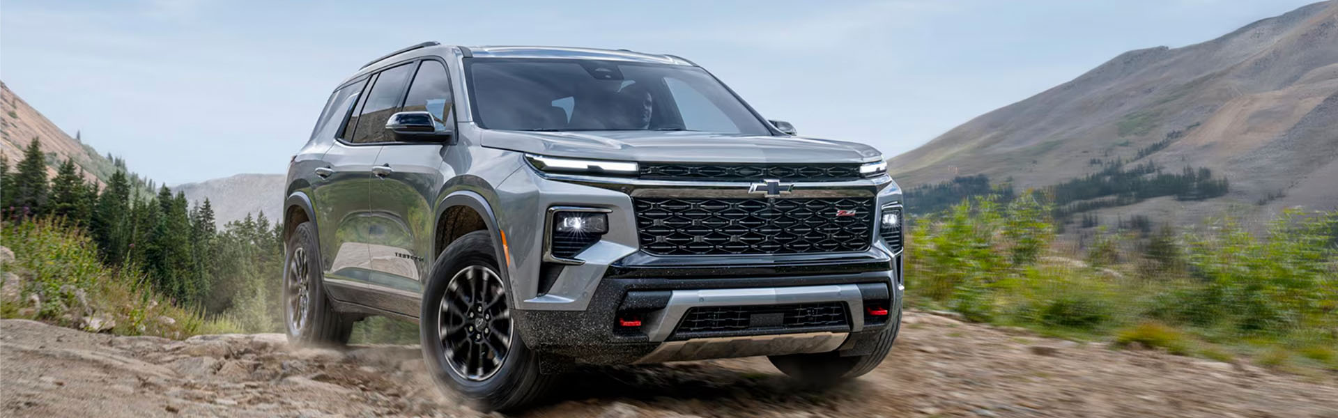 A silver 2026 Chevrolet Traverse SUV drives on a dirt path surrounded by mountains and trees.